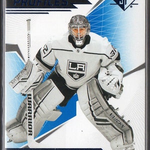 Può includere: Una carta da collezione di hockey con Jonathan Quick, un portiere dei Los Angeles Kings. La carta è bianca con accenti blu e argento. La carta ha il testo "Authentic Profiles" e "SP". Quick indossa una divisa da portiere bianca con il logo dei Los Angeles Kings sul davanti. È in posizione di portiere con la sua mazza nella mano sinistra e la mano destra sul suo guanto. La carta ha uno sfondo blu e argento con un motivo a griglia.
