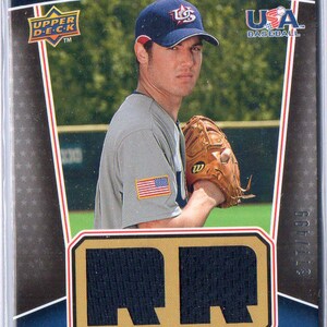 以下が含まれることがあります： 青い帽子に「USA」の文字が書かれた野球選手が写っている野球カード。選手は左袖に赤白青のアメリカ国旗のパッチが付いた白いユニフォームを着ている。カードは金色の縁取りがされており、上部には「USA Winning Materials」の文字が書かれている。選手の氏名「Robbie Ray」と「18U National Team」の文字がカードの下部に印刷されている。