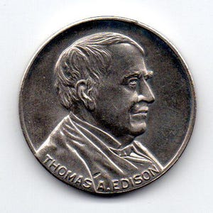 May include: A silver-colored coin featuring a profile of Thomas A. Edison. The coin has a detailed relief of Edison's face and the inscription "THOMAS A. EDISON" around the bottom edge. The coin has a circular shape and a metallic sheen.