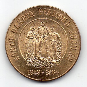May include: A gold-colored coin commemorating the North Dakota Diamond Jubilee. The coin features a depiction of three people standing together, with the text "North Dakota Diamond Jubilee 1889-1984" around the edge.