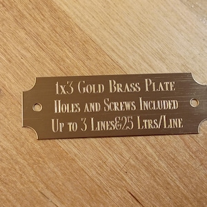 May include: A rectangular gold brass plate, 1x3 inches, with rounded corners and two holes. The text reads: "1x3 GOLD BRASS PLATE HOLES AND SCREWS INCLUDED UP TO 3 LINES&25 LTRS/LINE". Two gold screws are also visible.