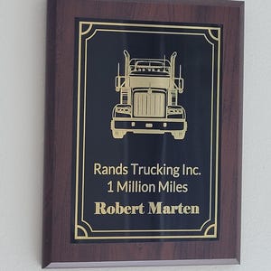 May include: A wooden plaque with a black plate featuring a gold outline of a semi-truck. The text reads "Rands Trucking Inc. 1 Million Miles Robert Marten."