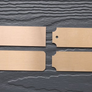 May include: Four blank metal nameplates, two with rounded corners and two with straight edges, on a dark wood background.