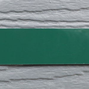 May include: A blank, rectangular sign with a dark green background. The sign is mounted on a gray, textured wall.
