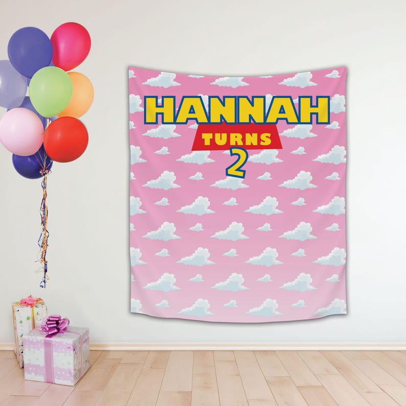 May include: Pink backdrop with a white cloud pattern. The backdrop has a yellow banner with the text "HANNAH TURNS 2" in blue letters.
