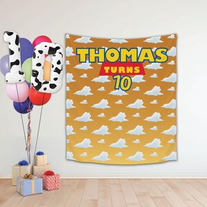 May include: A yellow and blue banner with the text "THOMAS TURNS 10" on a background of white clouds on a yellow sky.  The banner is hanging on a white wall with balloons in the foreground.