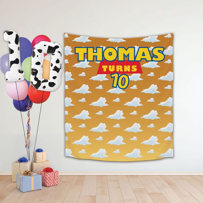 May include: A yellow and blue banner with the text "THOMAS TURNS 10" on a background of white clouds on a yellow sky.  The banner is hanging on a white wall with balloons in the foreground.