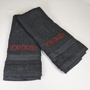 May include: Two black hand towels with the embroidered text "YOU DRIED" in red. The towels are folded and have a dark band at the bottom. The towels are made of a soft, absorbent material.