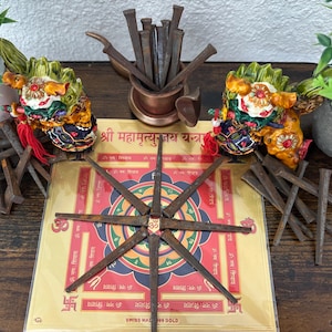 May include: A square Mahamrityunjay Yantra card with a central geometric mandala and Sanskrit text, surrounded by eight rusted iron nails arranged in a star pattern. Two colorful guardian lion figurines and a copper cup filled with nails are present.