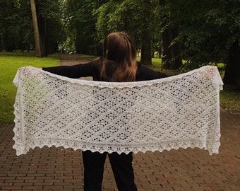 Pode incluir: Um xale branco de crochê com um delicado padrão de renda. O xale está drapeado sobre os ombros de uma pessoa em pé em um caminho de paralelepípedos em um parque.