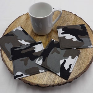 May include: Four square fabric coasters with a camouflage pattern in shades of brown, black, white, and gray. The coasters are on a wooden surface with a white mug.