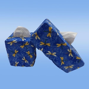 May include: Two tissue box covers in a blue fabric with a dragonfly pattern. The dragonfly design features yellow and white dragonflies with a swirling vine pattern. White tissues are visible in the openings.