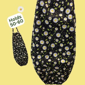 May include: A black fabric bag featuring a repeating pattern of white daisies with yellow centers and green stems. The bag has a gathered opening at the top and bottom. A white tag attached to the bag displays the text "Holds 50-60" in green.