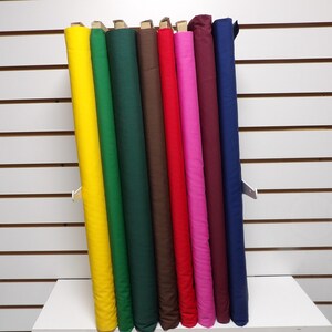 May include: A display of fabric rolls in various colors, including yellow, green, brown, red, pink, and blue. The rolls are neatly arranged and presented against a white background, showcasing the diverse color options available.