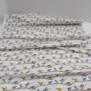 May include: White fabric with a repeating pattern of birds perched on brown branches. The birds are depicted in yellow and blue, with some nests visible. The fabric is folded, showing the pattern's continuity. Ideal for crafting or home decor projects.