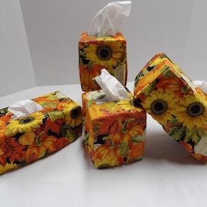 May include: Four tissue box covers with a fall theme. The covers are made of fabric with a pattern of sunflowers, pumpkins, and leaves in shades of orange, yellow, and green.