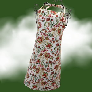 May include: A white apron featuring a repeating floral pattern in shades of red, orange, and green. The apron includes a bib top, a waist tie, and a large front pocket, displayed on a black wire mannequin form against a dark green background.