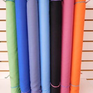 May include: A collection of fabric rolls in various colors, including green, blue, purple, black, pink, and orange. Each roll is secured with a rubber band. The rolls are displayed on a metal rod against a white wall.