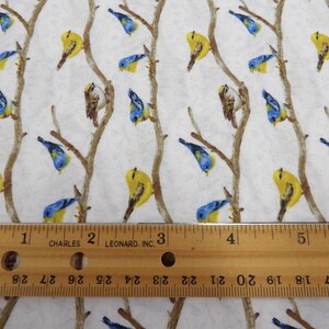 May include: White fabric with a repeating pattern of birds perched on brown branches. The birds are yellow, blue, and brown. A wooden ruler with inch measurements is in the foreground.