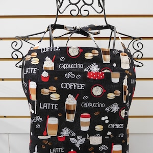 May include: Black apron with a coffee theme. The apron has a pocket and is decorated with images of coffee cups, coffee beans, and pastries. The text "LATTE", "COFFEE", "CAPPUCCINO", and "ESPRESSO" is printed on the apron.