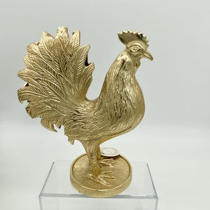 May include: A gold-colored rooster figurine with a textured, metallic finish. The rooster stands on a circular base with a radial pattern. The feathers on the tail and body are detailed with fine, etched lines, and the comb is visible.