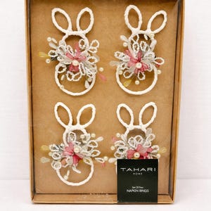 May include: A set of four bunny-shaped napkin rings. Each white ring is adorned with pink and white floral accents and faux pearls. The napkin rings are displayed in a brown cardboard box with the brand name "TAHARI HOME" on the label.