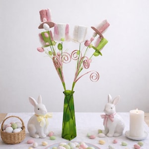 May include: Easter-themed still life featuring a green vase with decorative floral stems topped with miniature hats. White ceramic bunnies with bows, a basket of eggs, and a plate of pastel-colored candies complete the festive arrangement.