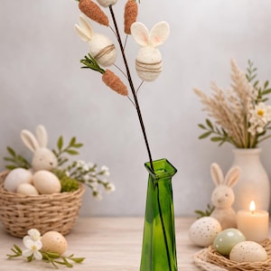 May include: A green glass vase holds a decorative Easter stem with felt bunny heads and carrot accents. The Easter-themed arrangement is surrounded by Easter eggs, a wicker basket, and a candle, creating a festive holiday display.