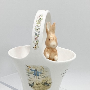 May include: A white ceramic basket with a floral design and a brown rabbit figurine peeking out of the top. The basket features a scene from the children's book "The Tale of Peter Rabbit" with a scarecrow and birds.
