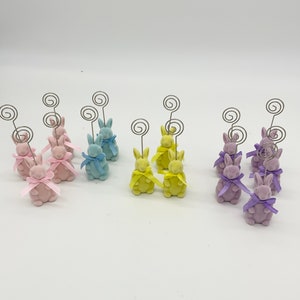 May include: A set of 10 bunny-shaped photo holders. The holders are made of ceramic and come in three colors: pink, blue, and yellow. Each bunny has a bow and a metal wire loop for holding photos.