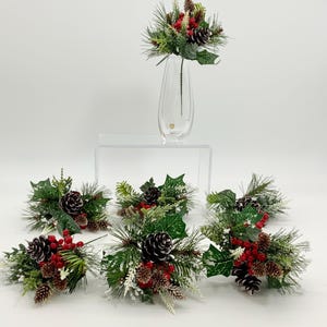 May include: A clear glass vase holds a festive arrangement of faux pine needles, red berries, and pine cones. Six additional arrangements of the same design are displayed around the vase, creating a holiday theme. The greenery is accented with white frost.