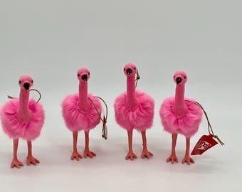 May include: Four pink flamingo ornaments with fluffy bodies, long necks, and slender legs. Each ornament has a small tag attached. The flamingos are standing on a white surface.