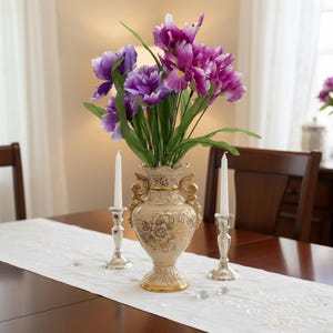 May include: A decorative centerpiece featuring a cream-colored vase with gold accents, filled with purple and pink flowers and green leaves. Two silver candlesticks with white candles sit on either side of the vase on a white table runner.