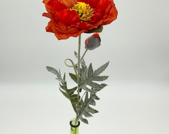 May include: A single red poppy flower with a green stem and leaves in a green glass vase. The flower has a yellow center and is in bloom.