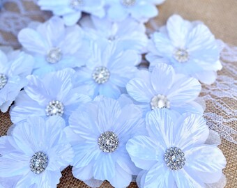 May include: A collection of white fabric flower embellishments with sparkling rhinestone centers. The flowers are arranged on a burlap surface with a delicate lace trim, creating a soft, elegant aesthetic. These floral accents are ideal for craft projects.
