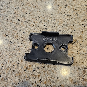 May include: A black plastic 3D printed mount with the word "ULAG" printed on it. The mount has two holes and a hexagonal cutout.