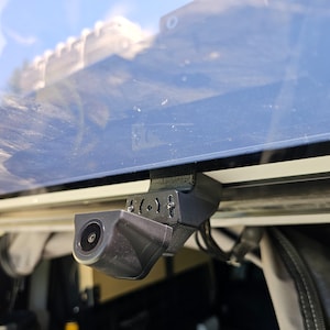 May include: A black rear-view camera mounted on a vehicle with a black strap. The camera is angled downwards and has a lens on the front.