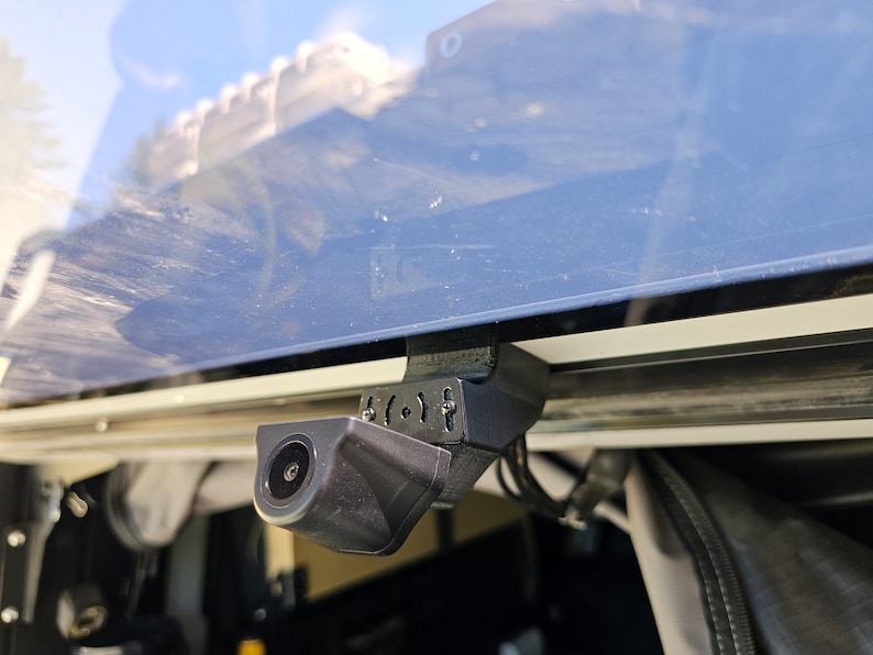 May include: A black rear-view camera mounted on a vehicle with a black strap. The camera is angled downwards and has a lens on the front.