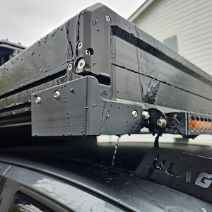 May include: A black roof top tent with a light bar mounted on the side. The tent is mounted on a black car. The tent is wet from rain.