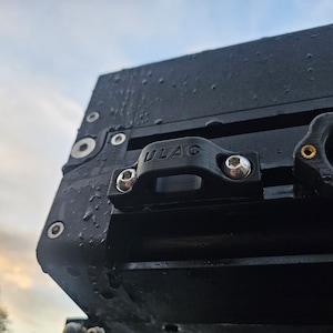 May include: A black metal box with a black plastic latch labeled "ULAG" and silver screws. The box is wet with water droplets.