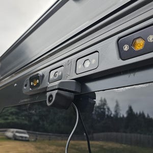 May include: A black rear-view camera mounted on the back of a vehicle. The camera is connected to a wire and has a lens facing backwards. There are three small lights on the vehicle's body above the camera.