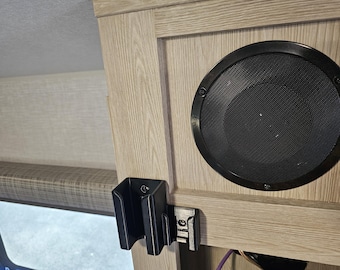 May include: A black speaker with a grill covering the speaker cone is mounted on a light wood surface. A black plastic holder is mounted to the left of the speaker.