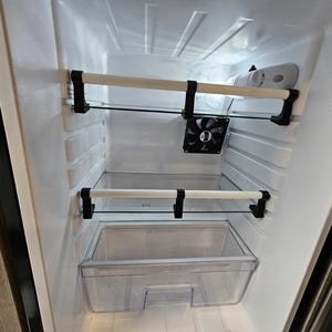 May include: Empty white refrigerator interior with two glass shelves and a clear plastic crisper drawer. The shelves are supported by black metal brackets. A small black fan is visible in the upper section of the refrigerator.