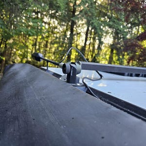 K&ouml;nnte beinhalten: Ein schwarzes Kamera- und Montagesystem, das auf einem Fahrzeugdach befestigt ist. Die Kamera ist klein und zylindrisch, mit einem nach vorne gerichteten Objektiv. Eine schwarze, rechteckige Stange erstreckt sich von der Halterung. Der Hintergrund zeigt einen Wald mit Sonnenlicht, das durch die B&auml;ume scheint.