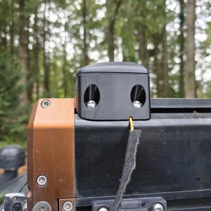 May include: A black, rectangular device with two circular openings, each containing a silver element. The device is mounted on a black and brown metal structure. A yellow wire and a black fabric strip extend from the device.