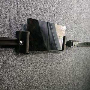 May include: A black tablet is secured in a black holder attached to a black rail system. The holder is mounted on a gray, textured surface. The tablet's screen reflects the surrounding environment.