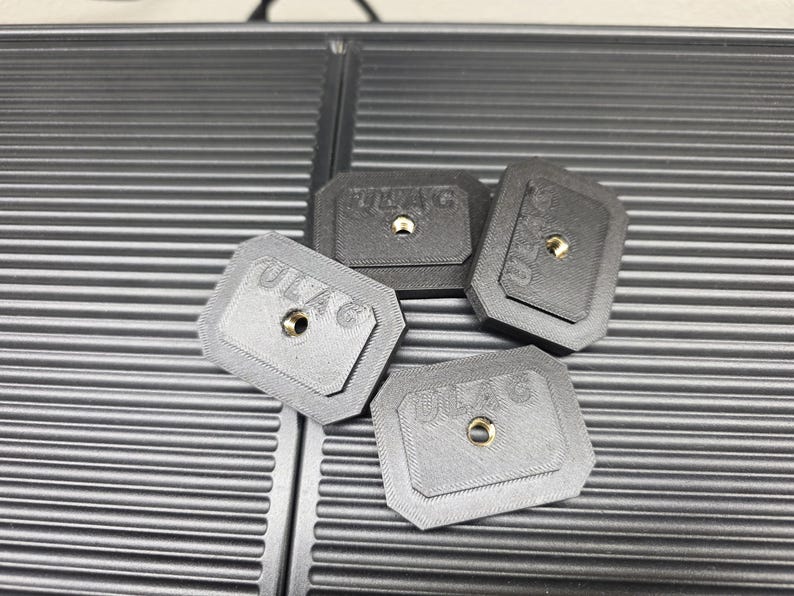 May include: Four small, rectangular, gray and black objects with the text "ULAG" and a central hole. The objects are arranged on a textured, dark gray surface.