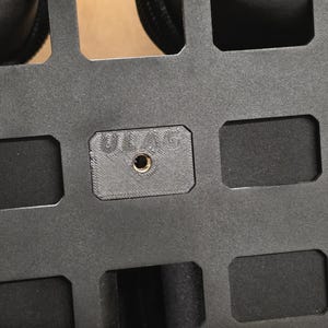 May include: Close-up of a black, rectangular panel with multiple rectangular cutouts. A small, square piece with the text "ULAG" and a central hole is visible. The panel appears to be made of a durable material.