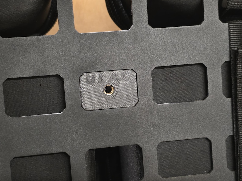 May include: Close-up of a black, rectangular panel with multiple rectangular cutouts. A small, square piece with the text "ULAG" and a central hole is visible. The panel appears to be made of a durable material.