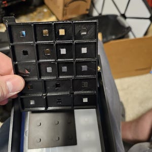 May include: A black, 3D-printed grid with sixteen square openings, held above a larger, partially visible structure. Some openings reveal metallic squares. The design appears to be a component of a larger electronic device or model.